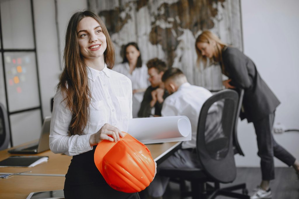 Smiling businesswoman with a hard hat in a modern office setting, engaging with colleagues.